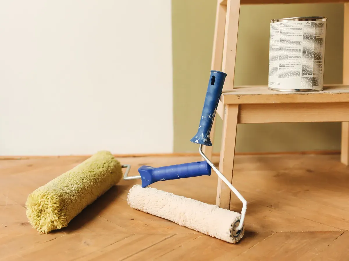 Paint rollers, a brush and an open can of paint on a workbench beside a freshly painted wall